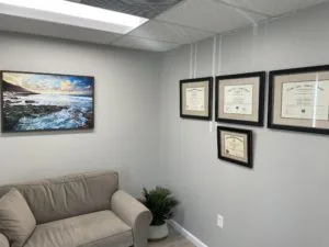 A wall poster of an ocean, an indoor plant beside the grey sofa and four certificates in a frame hanging on the wall